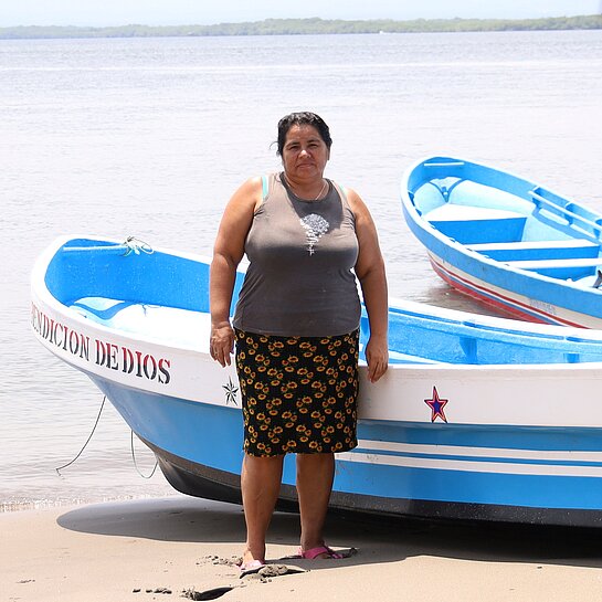 Eine Frau steht am Strand, im Hintergrund ist ein Fischerboot am Ufer zu sehen.