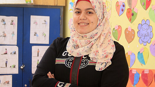 Eine junge Frau mit verschränkten Armen steht lächelnd in einem Klassenzimmer.