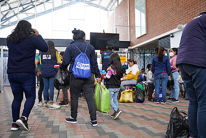 Mehrere Erwachsene und Kinder befinden sich in einem hellen, überdachten Raum. Frauen und jüngere Mädchen tragen Taschen und Beutel, teilweise mit Plan-Logo, manche von ihnen führen Kinder an den Händen.