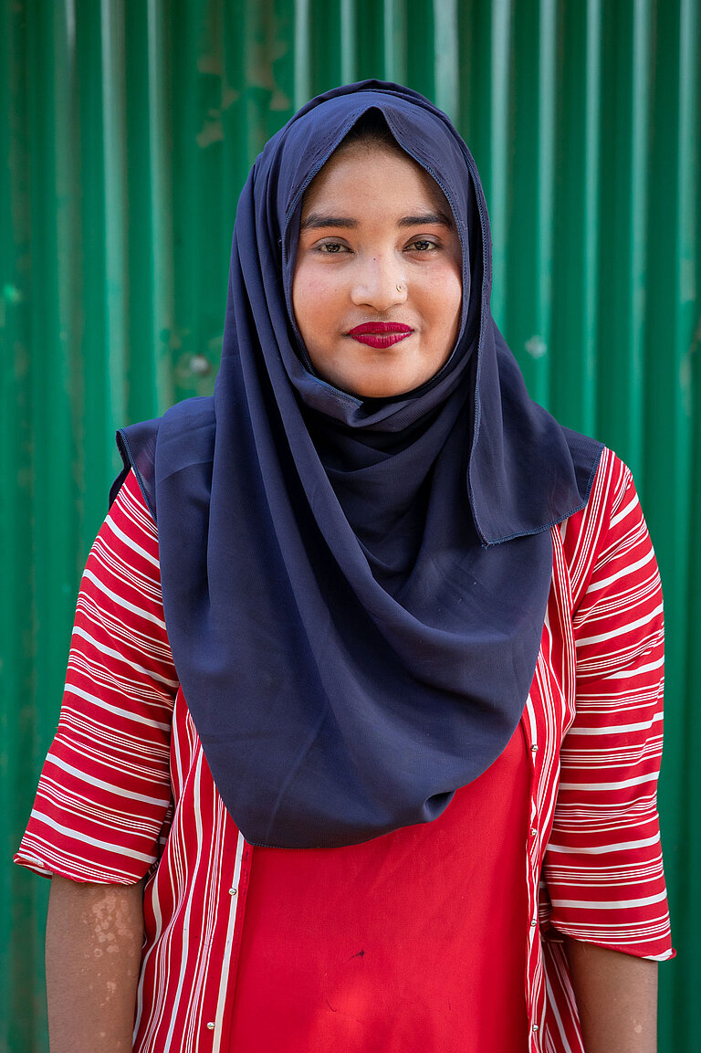 Jamila steht vor einer grünen Wand. Sie trägt eine rote Bluse und ein blaues Kopftuch und lächelt in die Kamera.