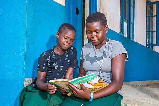 Vor einem neuen Grundschulgebäude in Uganda lesen zwei junge Mädchen zusammen in einem Buch
