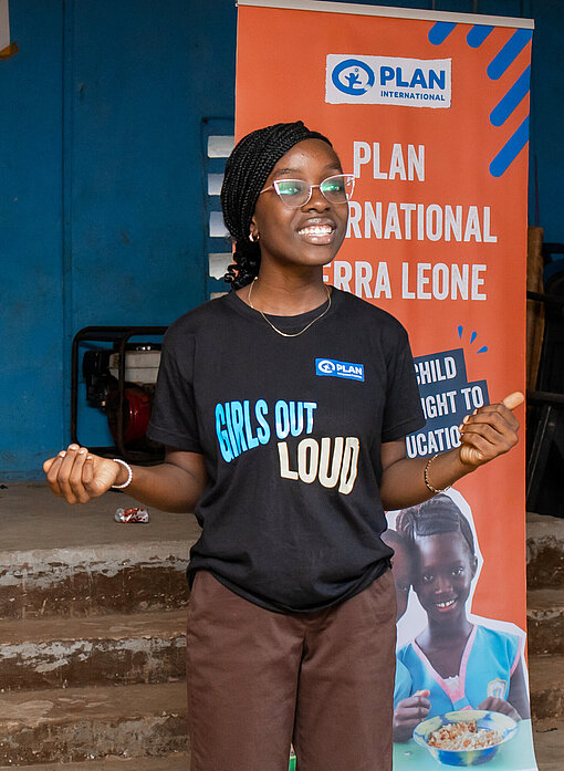Eine junge Frau steht vor einem Plan-Plakat und spricht angeregt. Sie trägt ein T-Shirt, auf dem "Girls Out Loud" geschrieben steht.