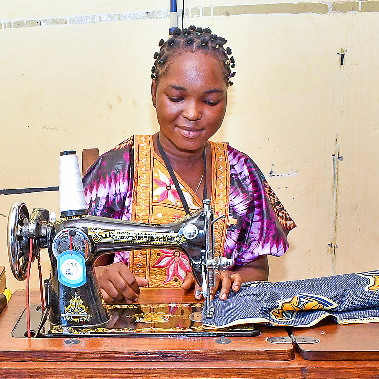 Eine junge Frau sitzt zufrieden an einer Nähmaschine und vernäht einen bunten Stoff
