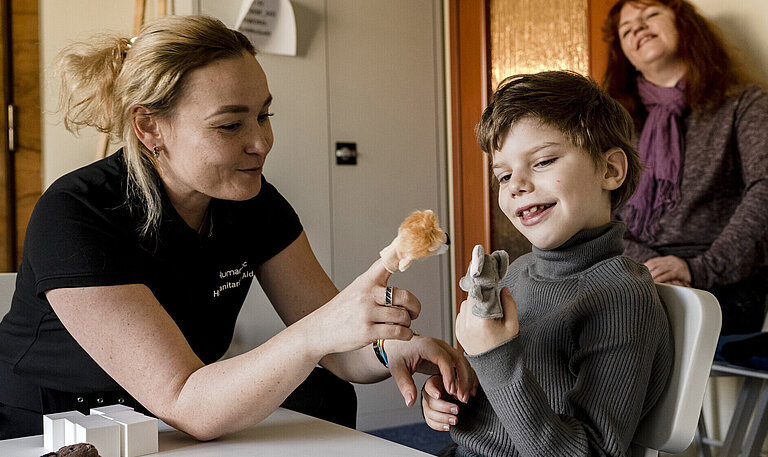 Eine Frau sitzt neben einem Jungen an einem Tisch. Die beiden spielen mit Fingerpuppen und lachen