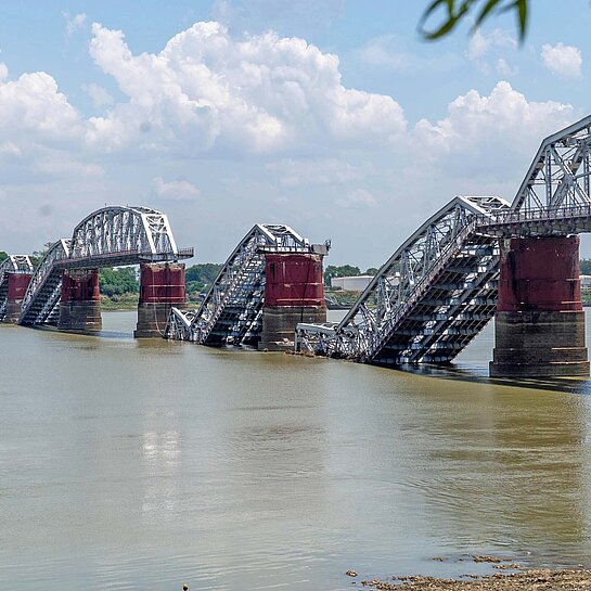 Blick auf eine Eisenbrücke, deren Bögen zerstört halb abgestürzt im Wasser eines Flusses liegen