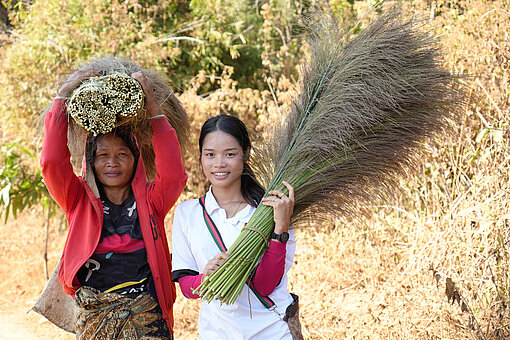 Zwei Frauen tragen Büschel von Besengras. Im Hintergrund ist ein Feld zu sehen.
