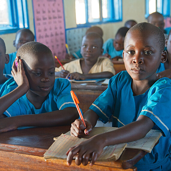 Mehrere Kinder sitzen in einer Klassensituation an Holzbänken. Sie tragen blaue Schuluniformen. Ein Kind im Vordergrund schreibt konzentriert in sein Heft, während ein anderes Kind sich über das Heft beugt. 