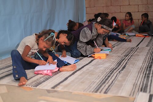 Mehrere Kinder sitzen auf Teppichen am Boden und malen oder schreiben in Hefte. Vor ihnen liegen Stifte und kleine Federmäppchen, im Hintergrund sitzen weitere Kinder.