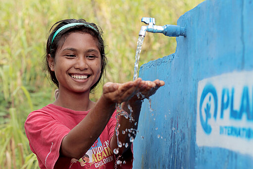Auch Eci holt Wasser jetzt vom neu eingerichteten Brunnen im Dorf Ein Mädchen mit pinkem T-Shirt steht lachend an einem Wasserhahn, aus dem Wasser sprudelt