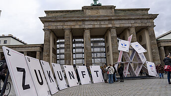 Vor dem Brandenburger Tor steht ein überdimensional großes Kartenhaus, das in sich zusammenbricht