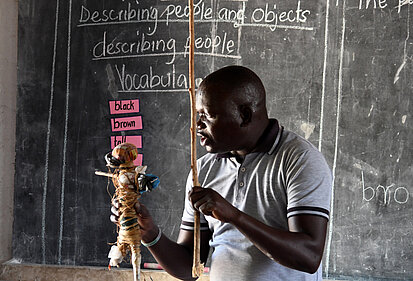 Ein erwachsener Mann steht vor einer Kreidetafel im Klassenraum und hält eine handgemachte Puppe und einen Stock in den Händen.