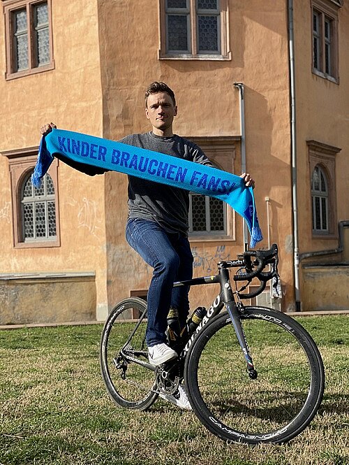 Martin sitzt auf dem Fahrrad und hält einen Schal mit der Aufschrift Kinder brauchen Fans hoch