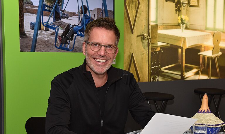 Schauspieler Tim Bergmann sitzt an einem Tisch und hält ein Blatt Papier in der Hand
