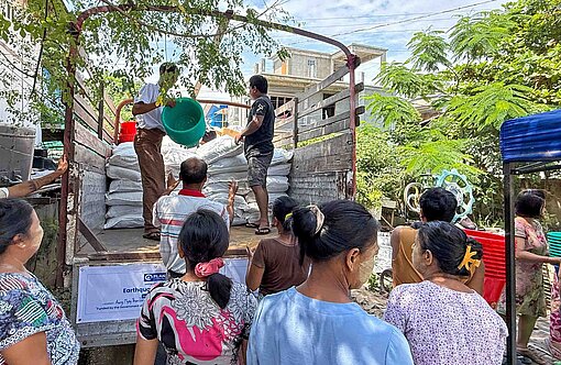 Frauen und Männer stehen vor einem Lastwagen, der Säcke mit Reis und andere Hilfsgüter anliefert