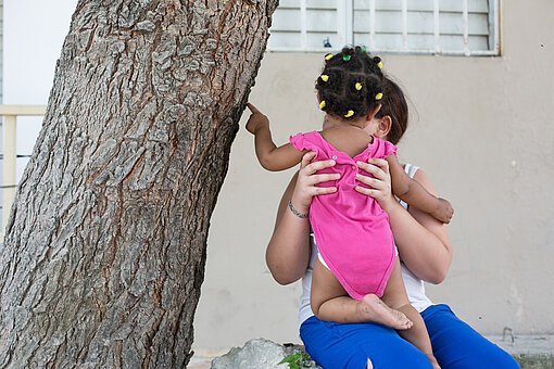 Bivian mit Schwester Ein Mädchen hat ein Kleinkind auf dem Arm, das Kleinkind ist von hinten zu sehen und verdeckt das Gesicht des Mädchens