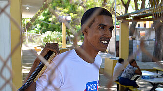 Carlos ist durch einen Maschendrahtzaun hindurch fotografiert. Er lächelt und schaut in die Ferne.