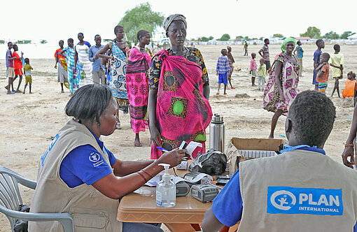 Plan-Teams verteilen in Zusammenarbeit mit dem Welternährungsprogramm der Vereinten Nationen (WFP) Nahrungsmittelhilfe an Familien im südsudanesischen Pibor Zwei Personen mit Plan-Jacken sitzen an einem Tisch