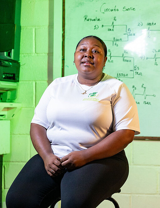 Eine junge Frau sitzt auf einem Hocker vor einer Tafel mit elektronischen Schaltzeichnungen; der Raum ist als Technik‑ und Lernumgebung mit Geräten und Unterrichtsmaterial erkennbar.