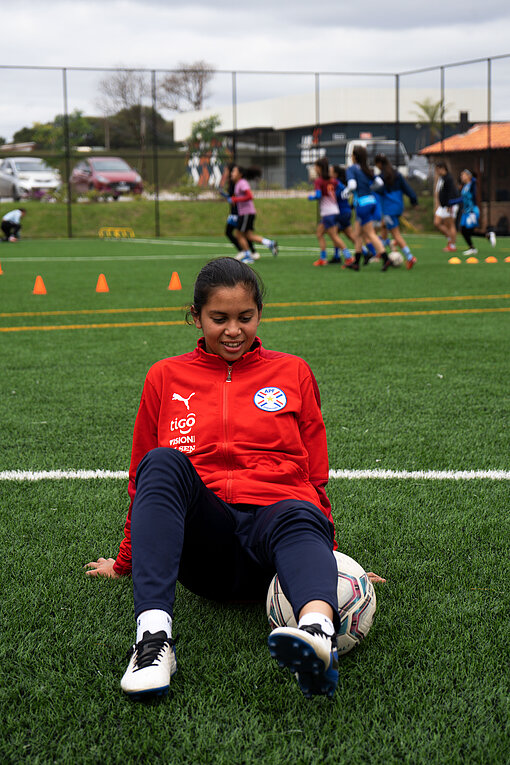 Mädchen in rotem Trainingsanzug sitzt auf dem Fußballrasen und wärmt sich mit einem Fußball auf