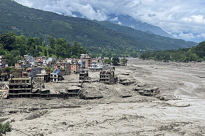 Ein vom Schlamm überschwemmtes Dorf in Nepal. Viele Häuser sind teilweise zerstört und von dicken Schlammschichten bedeckt. Im Hintergrund sind grüne bewaldete Berge und ein bewölkter Himmel zu sehen.