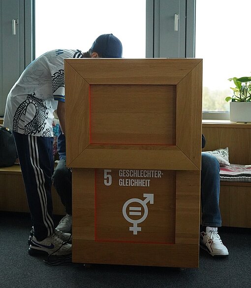 Zwei Schüler sitzen hinter einer großen Holzkiste, die aufgeklappt ist. Ein Schüler steht neben der Kiste und beugt sich darüber. Auf er Kiste steht: 5 Geschlechtergleichheit