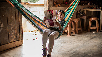 Eine junge Frau sitzt in einer Hängematte und hält ein Prospekt in der Hand. Sie lächelt in die Kamera.