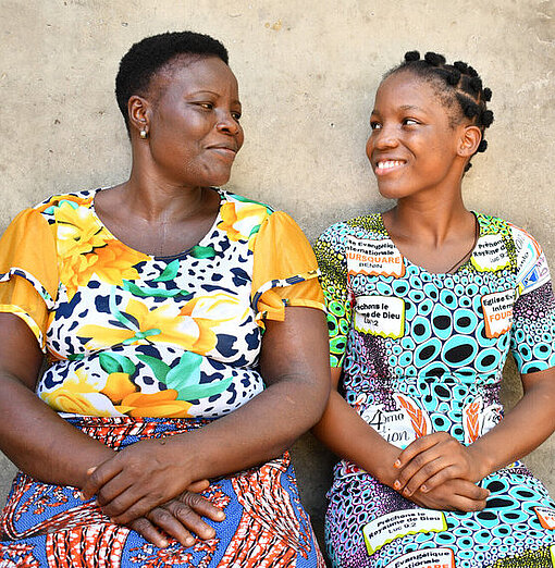 Monique und ihre Mutter sitzen auf einer Bank vor ihrem Haus Eine Mutter und Tochter aus Benin in bunter Kleidung sitzen auf einer Bank vor ihrem Haus