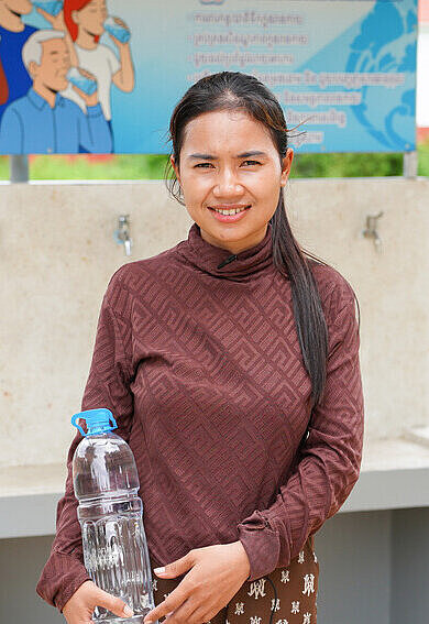 Eine Frau steht vor einem Wasserspender und hält ihre Wasserflasche mit sauberem Trinkwasser.