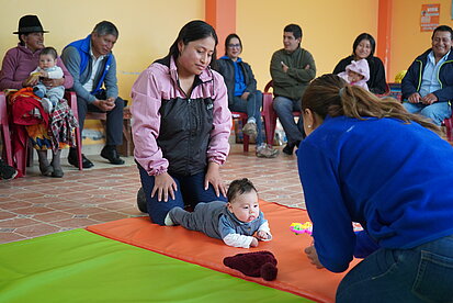 Im Vordergrund befindet sich eine junge Frau, die sich auf einem bunten Spielteppich neben einem Kleinkind kniet, das auf dem Bauch liegt und nach vorne schaut. Eine weitere junge Frau beschäftigt sich aufmerksam mit dem Kind, vermutlich im Rahmen einer frühen Förderung oder Beratung. 