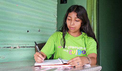 Eine junge Frau mit grünem T-Shirt sitzt am Schreibtisch und notiert etwas