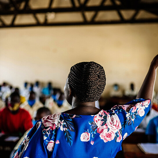 Eine Lehrerin mit Flechtfrisur steht mit dem Rücken zur Kamera einer Klasse zugewandt und hebt den Arm.