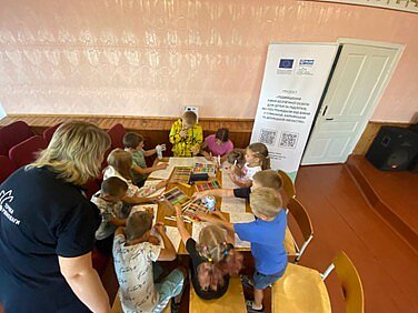 Kinder sitzen an einem Tisch und malen, eine Betreuungsperson steht neben dem Tisch und interagiert mit den Kindern