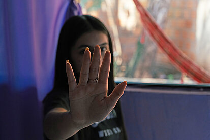 Eine junge Frau hält ihre rechte Hand in die Kamera und verdeckt durch die Geste ihr Gesicht. Es kann als Abwehr- beziehungsweise Stopp-Geste interpretiert werden. 