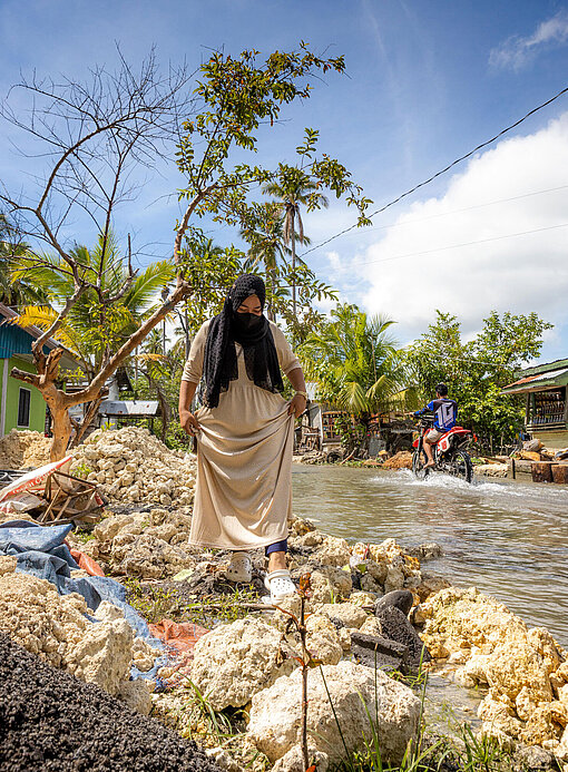 Farhana rafft ihr cremefarbenes Kleid hoch, um neben der überfluteten Straße über Gesteinsbrocken laufen zu können.
