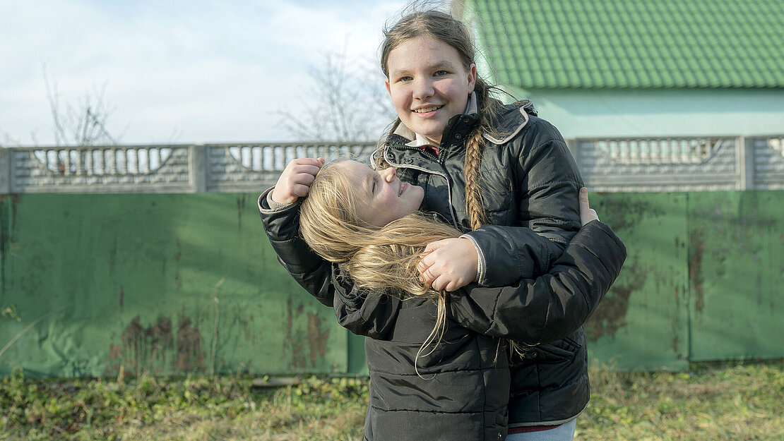Zwei Mädchen stehen draußen auf einer Wiese, umarmen sich und lachen dabei