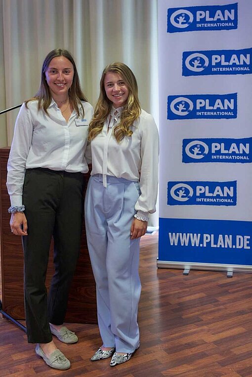Paula und Kim haben den Jugendbeirat bei der Mitgliederversammlung in Dresden vertreten. Zwei junge Frauen stehen in schicker Kleidung neben einem Plan International-Banner.