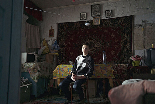 Ein Jugendlicher sitzt mitten in einer Küche auf einem Stuhl und schaut in die Kamera