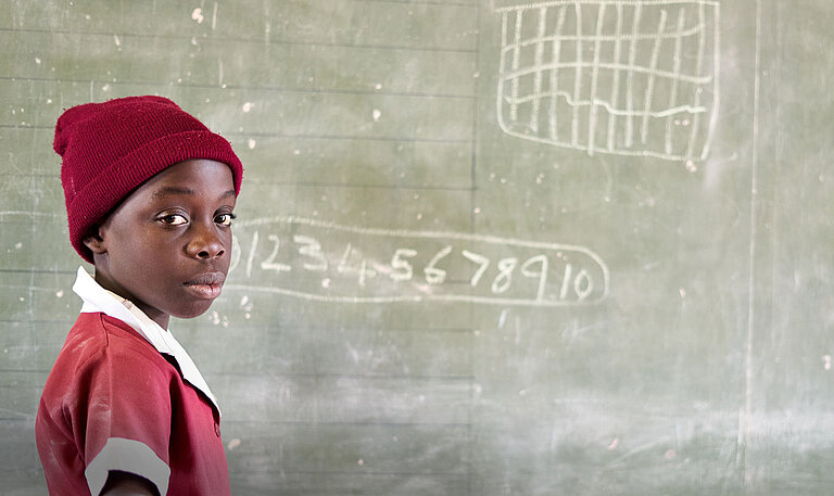 Ein Mädchen in rotem T-Shirt steht an einer Schultafel, auf der Zahlen mit Kreide notiert sind