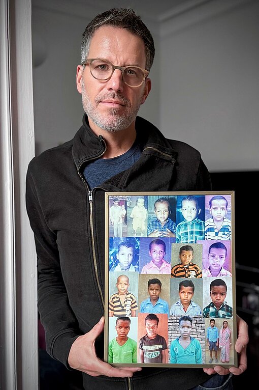 Schauspieler Tim Bergmann steht an einen Türrahmen gelehnt und zeigt eine gerahmte Collage mit Portraitfotos seiner Patenkinder 