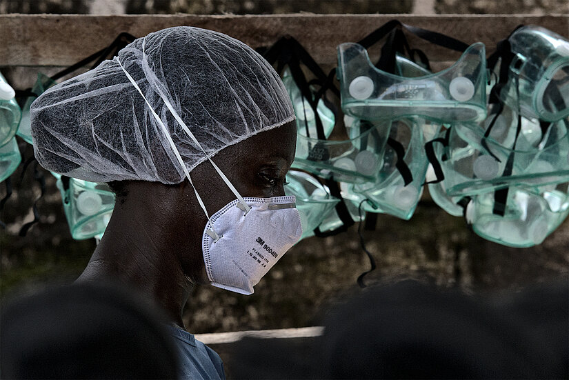 Eine Person trägt eine Schutzmaske und ein Haarnetz. Im Hintergrund hängen mehrere Schutzbrillen an einem Gestell. Die Szene wirkt wie ein Bereich mit Schutzkleidung, vermutlich während eines Ebola-Einsatzes.