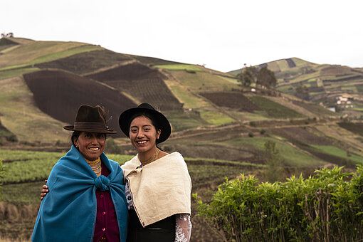 Eine junge und eine ältere Frau in indigener Kleidung vor hügeliger Landschaft