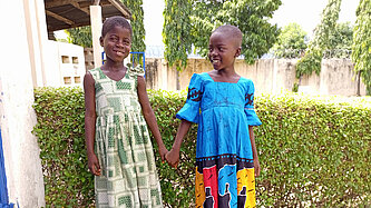  Zwei Mädchen in bunten Kleidern halten sich an den Händen und lächeln – Aufnahme in einer Schule in Togo.