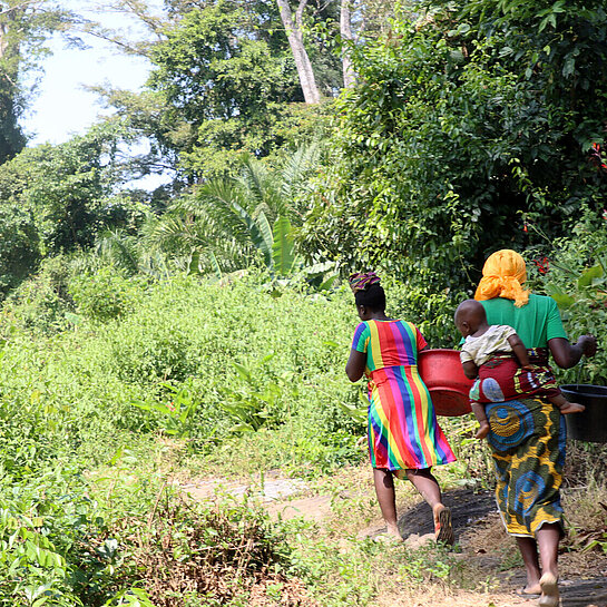 Zwei liberische Frauen laufen mit Wassereimern auf dem Kopf durch den Wald. Eine von ihnen trägt ein Kind auf dem Rücken