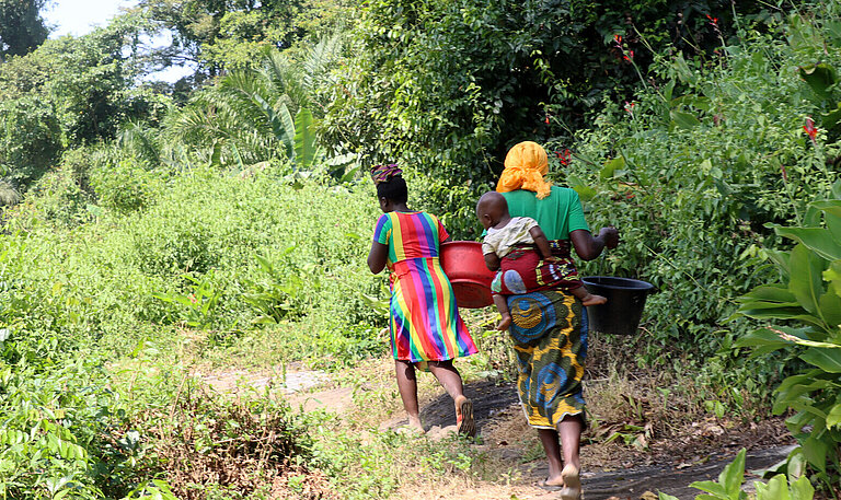 Zwei liberische Frauen laufen mit Wassereimern auf dem Kopf durch den Wald. Eine von ihnen trägt ein Kind auf dem Rücken