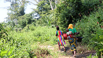 Zwei liberische Frauen laufen mit Wassereimern auf dem Kopf durch den Wald. Eine von ihnen trägt ein Kind auf dem Rücken