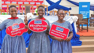 Drei Mädchen in Schuluniform halten rote Schilder mit den englischen Aufschriften „End Period Poverty“, „End Period Stigma“ und „Gender Equality Reusable Pad“ und lächeln in die Kamera. Im Hintergrund sind weitere Menschen und ein Bühnendekor zu sehen.