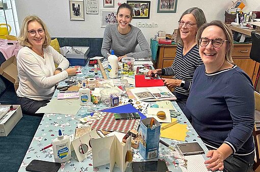 In einer Küchenstube sitzen vier Frauen über einen Tisch gebeugt und basteln mit Papier und Schere