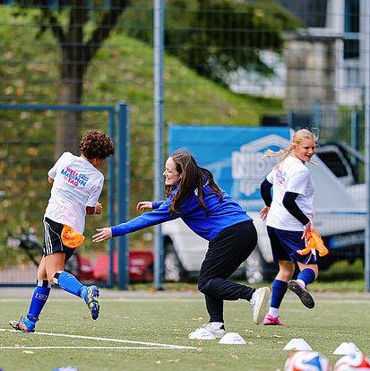 Zwei Mädchen in weißen Shirts mit der Aufschrift "Weltmädchentag" spielen mit einer Frau fangen auf einem Fußballplatz.