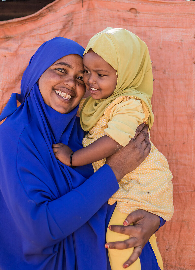 Cawo hält ihre einjährige Tochter im Arm, Cawo lächelt und ihre Tochter schaut sie lächelnd an.
