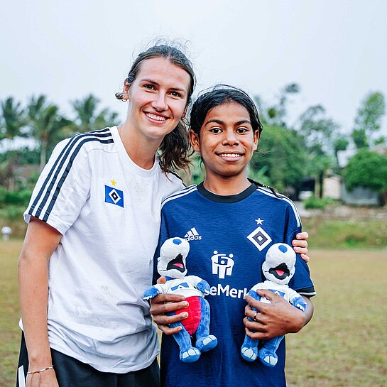 Eine junge Frau mit weißem HSV-Trikot steht zusammen mit einem dunkelhaarigen kleinen Mädchen in dunkelblauem HSV-Trikot zusammen.